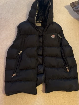 Svart dunväst från Moncler - Snygg svart dunväst från Moncler med huva och dragkedjor framtill. Västen har två stora fickor med dragkedja och Moncler-logga på bröstet. Insidan har Moncler-tryck längs kanterna och quiltad design. Perfekt för lager på lager och streetwear. Storlek XXL (herr). Självklart äkta samt har en unik QR kod som kan intyga detta. I mycket fint skick, väl omhändertagen samt sparsamt använd.  
