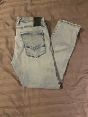 Ljusblå Replay Hyperflex jeans - Säljer ett par ljusblå Replay Hyperflex jeans med klassisk femficksdesign och snygga kontrastsömmar. Jeansen har en rak passform och är tillverkade i stretchigt material för extra komfort. Bakfickan har en unik broderad detalj. Har en reva i skrevet, men syns knappt när man har på sig dem
