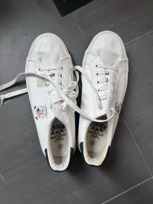 Vita sneakers från Vinskon Polo Club - Klassiska vita sneakers från Vinskon Polo Club med svarta detaljer på hälen och logga på sidan. Skorna har lågt skaft, vita snören och är tillverkade i ett slätt syntetmaterial. Perfekta för en clean och stilren look.