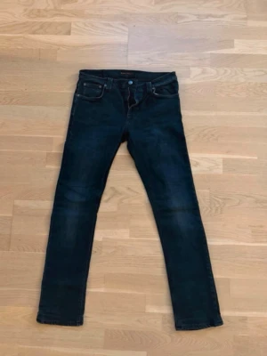 Mörkgrå slim fit jeans från Nudie - Säljer ett par mörkgrå/svart slim fit jeans från Nudie Jeans med klassiska kontrastsömmar och broderade bakfickor i blå färg och är tillverkade i mjuk denim med lätt tvättad look.