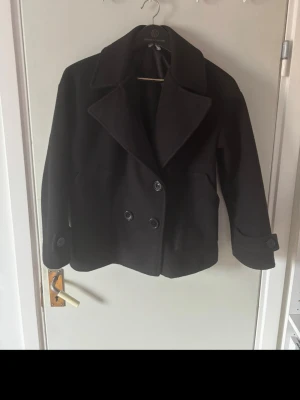 Svart kort rockfrån Design By Si - Stilren svart rock med dubbelknäppning och bred krage. Modellen är kortare och har stora knappar samt uppvikta ärmslut. Perfekt för dig som vill ha en klassisk men modern look. XS/S