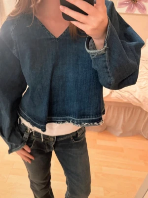 Jeanstopp/denim topp - Mörkblå croppad jeanstopp med v-ringning och långa ärmar. Blusen har råa, fransiga kanter nertill och är tillverkad i bomull. 