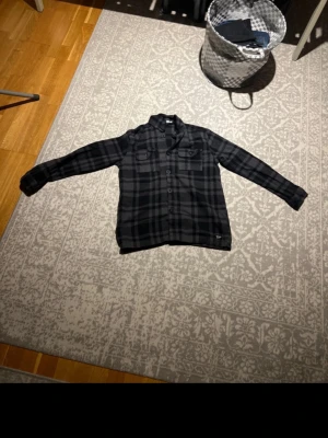 Mörkgrå rutig skjorta - Säljer en mörkgrå rutig skjorta med knappar framtill och bröstficka. Skjortan har lång ärm och klassisk krage. Perfekt för dig som gillar en avslappnad men ändå stilren look.