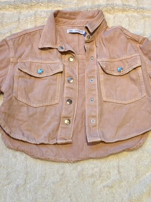 Kort rosa jeansjacka från Zara - Trendig kort jeansjacka i ljusrosa från Zara med två bröstfickor och snygga silverknappar. Jackan har klassisk krage och avslappnad passform, perfekt för att lyfta din outfit med en cool touch. Materialet är mjukt jeans och jackan har korta ärmar.