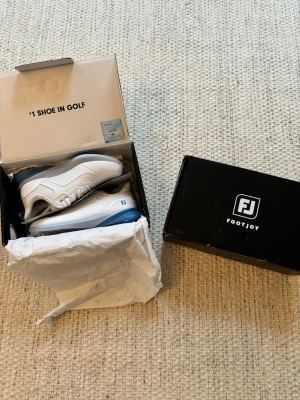 Vita golfskor från FootJoy - Snygga vita golfskor från FootJoy med blå sula och detaljer. Skorna har en sportig design med ventilerande mesh och slitstarka syntetmaterial. Perfekta för golfbanan med greppvänlig yttersula och klassisk FootJoy-logga på sidan.