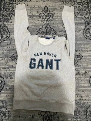 Grå sweatshirt från GANT - Snygg grå sweatshirt från GANT med trycket 'NEW HAVEN GANT' i mörkblått framtill. Tröjan har rund halsringning med randig detalj och mjuk, borstad insida för extra komfort. Perfekt för en avslappnad och sportig stil.