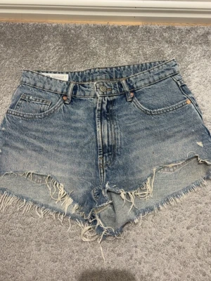 Blå Zara jeansshorts med fransar - Säljer ett par klassiska blå jeansshorts från Zara med mid waist midja och råa, fransiga kanter. Shortsen har fem fickor, knappgylf och slitna detaljer för en avslappnad look. Perfekta till sommaren och festivaler. Sitter som S🫰🏼
