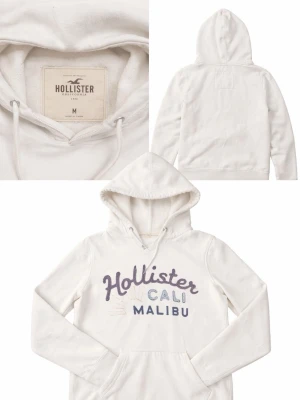 Vit hoodie från Hollister med tryck - Snygg vit hoodie från Hollister med stor magficka huva. Framsidan har ett broderat tryck med texten 'Hollister CALI MALIBU' i lila och blått. Tillverkad i mjuk bomullsblandning, perfekt för chill dagar. Klassisk passform och ribbade muddar.