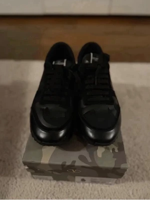 Svarta Valentino sneakers med camo - Snygga svarta sneakers från Valentino med diskret camo-mönster på ovandelen. Skorna har klassisk låg siluett, svarta skosnören och detaljer i läder och textil. Perfekta för dig som vill ha en exklusiv och stilren look.