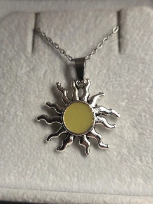Halsband med solhänge som lyser i mörkret - Unikt halsband med ett solformat hänge i rostfritt stål med en gul mitt som lyser blått/grönt i mörkret. Kedjan är tunn och stilren, och hänget har en cool, vågig design som verkligen sticker ut. Perfekt för dig som gillar accessoarer med lite extra edge och glow. Kedjans längd är 45 cm och hänget ca 2,5 - 3 cm. Vanligt dagsljus en kort stund räcker för att hänget ska ändra färg när man kommer in i en mörkare omgivning.