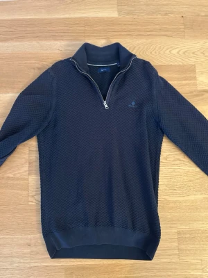 Blå halvzip tröja  - Snygg mörkblå tröja från GANT med halvzip och diskret strukturmönster. Tröjan har hög krage, långa ärmar och ribbade muddar. Perfekt för en stilren och avslappnad look.