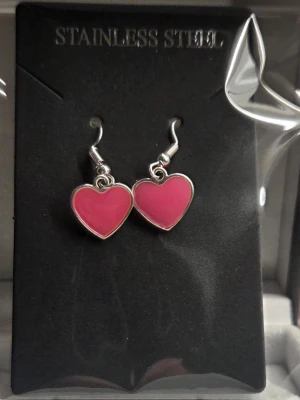Rosa hjärtformade örhängen - Säljer ett par söta hjärtformade örhängen i rosa med silverfärgad kant. Örhängena är tillverkade i stainless steel och lyser i mörkret med en lila nyans. Perfekta för dig som vill sticka ut lite extra!