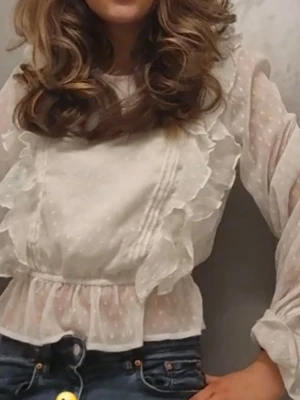 Vit volangblus från NA-KD - Supersöt vit blus från NA-KD med volangdetaljer längs bröstet och axlarna.Sjölvklart tvättas sminkfläckarna bort innan postning.Blusen är gjord i ett tunt, transparent tyg med diskret prickmönster och har resår i midjan för en snygg passform. Långa ärmar med resår vid handlederna ger en romantisk vibe.