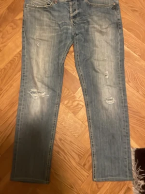 Ljusblå slitna jeans med raka ben - Säljer ett par ljusblå jeans med raka ben och slitningar på både fram- och baksida. Jeansen har klassiska fem fickor och synliga slitdetaljer på låren och knäna för en trendig look. Materialet är denim och passformen är avslappnad. Har sytt under byxorna men märks inte vid användning 