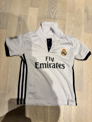 Real Madrid Ronaldo matchtröja - Vit Real Madrid matchtröja med svarta detaljer, korta ärmar och tryckta loggor. På framsidan står Fly Emirates och klubbmärket, på baksidan står Ronaldo och nummer 7. Tillverkad i funktionsmaterial, perfekt för fotbollsträning.