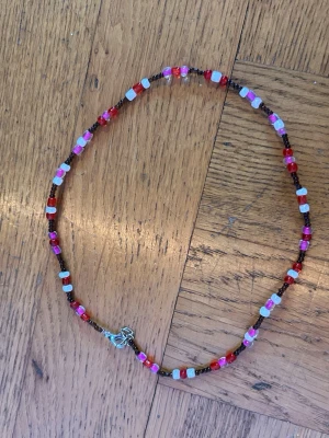 Halsband med färgglada pärlor - Halsband med små pärlor i rosa, rött, vitt och brunt. Pärlorna är i olika former och ger ett lekfullt och trendigt intryck. Halsbandet har ett enkelt metallspänne och är gjort av glas- och plastpärlor.