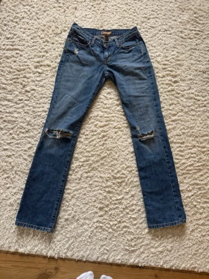 Blå lågmidjade bootcut jeans med slitningar från Abercrombie & Fitch - Säljer ett par lågmidjade bootcut jeans med utsvängda ben och slitningar på båda knäna samt en mindre reva vid fickan. Klassisk femficksmodell med knappgylf och coolt tvättad denim. Perfekta för en avslappnad och trendig look. Jag säljer de på grund av att de är för långa för mig. Midjemått: 36 cm Innerbenslängd: 85 cm💞
