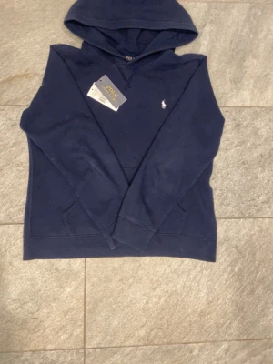 Marinblå hoodie från Polo Ralph Lauren - Snygg marinblå hoodie från Polo Ralph Lauren med klassisk huva och känguruficka. Ikoniska vita logon broderad på bröstet. Tillverkad i mjukt material som känns skönt mot huden. Perfekt för en avslappnad och stilren look.