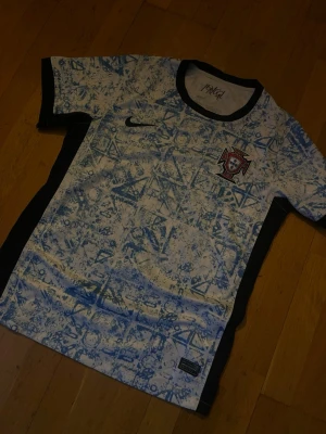 Portugal Nike fotbollströja blåvit - Portugal fotbollströja från Nike i vitt med blått grafiskt mönster. Tröjan har svarta detaljer vid krage och ärmslut samt Portugals landslagsemblem på bröstet. Perfekt för fotbollsträning eller att bära som supporter.