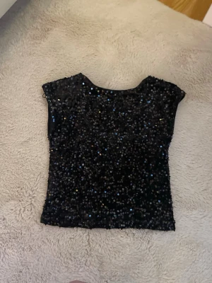 Svart paljettopp med öppen rygg - Glittrig svart topp täckt med paljetter. Modellen är croppad och ärmlös, perfekt för att sticka ut. Ingen synlig logga eller märke. Materialet känns som polyester.