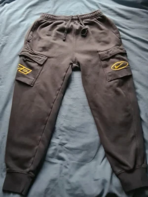 Mörkgrå nike tracksuit med gula detaljer. Tröja size:S Byxor size:M - Säljer ett par mörkgrå nike tracksuit med resår i midjan och snörning. Byxorna har stora fickor på sidorna med gula broderade detaljer, bland annat siffran 72 och en grafisk logga. Mjukt material, perfekta för en avslappnad streetwear-look..