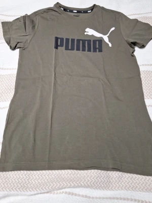 Olivgrön Puma t-shirt med logga - Snygg olivgrön t-shirt från Puma med stor svart och vit logga på bröstet. Klassisk rund halsringning och korta ärmar. Tillverkad i mjuk bomull som känns skön mot huden. Perfekt för en avslappnad och sportig stil. St 164