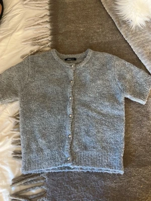 Ljusgrå kortärmad kofta Gina Tricot - Mysig ljusgrå kofta från Gina Tricot med korta ärmar och rund halsringning. Koftan har en fluffig, mjuk struktur och stängs med silvriga knappar framtill. Perfekt för lager på lager och en chill look.