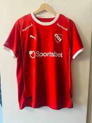 Independientes fotbollströja  - Snygg röd fotbollströja från Puma med vita detaljer och klubbmärke för Club Atlético Independiente. Tröjan har tryckta sponsorer som Sportsbet, Kanji och Pardo samt vita muddar vid ärmar och hals. Perfekt för dig som älskar fotboll och vill sticka ut på planen.