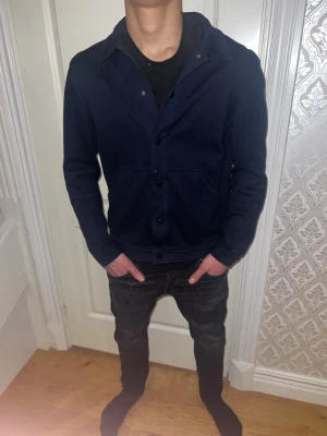 Mörkblå overshirt med knappar - Snygg mörkblå overshirt med klassisk krage och knäppning framtill. Två stora fickor framtill och raka ärmar. Overshirten är gjord i ett kraftigare tyg som ger en stilren look. Perfekt att bära över en t-shirt för en avslappnad men trendig stil.