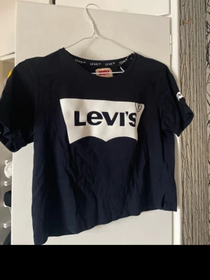 Svart croppad Levi's t-shirt - Snygg svart croppad t-shirt från Levi's med stort vitt logotryck på bröstet. Klassisk rund halsringning och korta ärmar. Perfekt för en avslappnad och trendig look. Tillverkad i mjuk bomull för skön känsla.