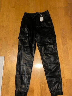 Svarta cargobyxor i fuskskinn - Säljer ett par svarta cargobyxor i fuskskinn med resår vid benslut och stora fickor på sidorna. Byxorna har en modern, avslappnad passform och cool streetwear-vibe. Perfekta för dig som vill sticka ut med en edgy look.