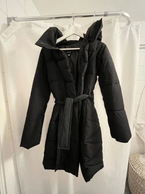 Svart lång vinterjacka storlek xs från Veromoda - Snygg svart lång pufferjacka med huva och bälte i midjan. Jackan har quiltad design, långa ärmar och en markerad midja för en clean siluett. Stor luva/krage. Storlek xs. Knappt använd. 