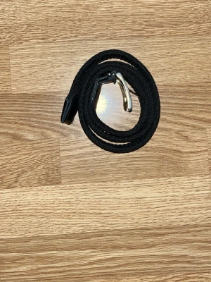 Grisch Svart flätat bälte med silverfärgat spänne - Stilrent svart flätat bälte med silverfärgat metallspänne och detaljer i skinn. Perfekt accessoar för att lyfta din outfit med en klassisk touch. Bältet är flexibelt och passar till många olika stilar.