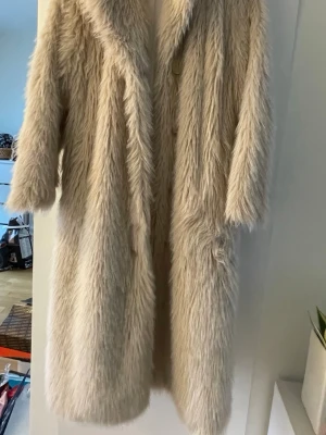 Lång beige fuskpälsjacka - size 36 men går till 38. Säljer en lång, fluffig beige fuskpälsjacka med bred krage och dolda knappar framtill. Jackan har en oversized passform och når ner mot knäna. Perfekt för dig som vill ha en trendig och varm vinterlook.