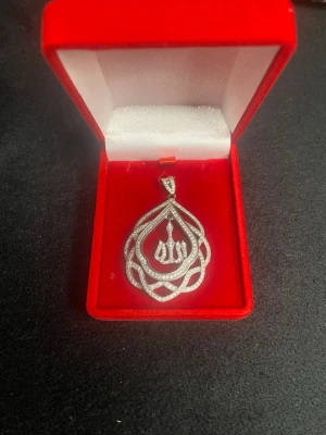 Silver halsband med gnistrande stenar - Unikt halsband med ett stort, droppformat hänge i silverfärg, täckt av små gnistrande stenar. Hänget har ett intrikat mönster och arabisk kalligrafi i mitten. Perfekt för dig som gillar detaljerade smycken med bling.