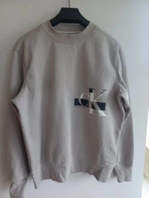Beige sweatshirt från Calvin Klein - Snygg beige sweatshirt från Calvin Klein med stort CK-tryck i vitt och svart på framsidan. Tröjan har rund halsringning, ribbade muddar vid ärmslut och nederkant samt en avslappnad passform. Perfekt för en casual och trendig look.