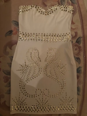 Vit axelbandslös klänning med nitar - Snygg vit axelbandslös kortklänning dekorerad med guldfärgade nitar i mönster av två svanar framtill. Klänningen har hjärtformad urringning och markerad midja. Perfekt för dig som vill sticka ut med coola detaljer och unik design.