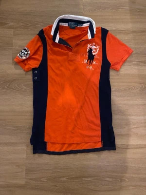 Orange pikétröja från Polo Ralph Lauren - Snygg orange pikétröja från Polo Ralph Lauren med vita och marinblå detaljer. Tröjan har klassisk krage, tryckt logga på bröstet och patch på ärmen. Tillverkad i bomull med custom fit-passform. Perfekt för en sportig och avslappnad stil.