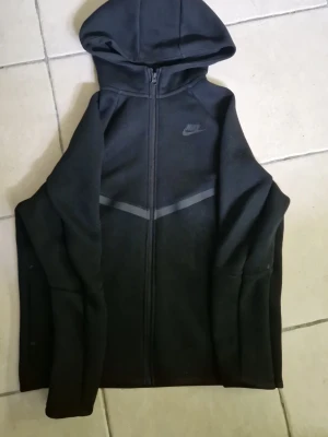 Svart Nike hoodie med dragkedja - Svart hoodie från Nike med hel dragkedja och huva. Snygga grå detaljer i form av V-mönster över bröstet och en diskret Nike-logga på vänster sida. Tillverkad i mjukt material, perfekt för en sportig och avslappnad stil.