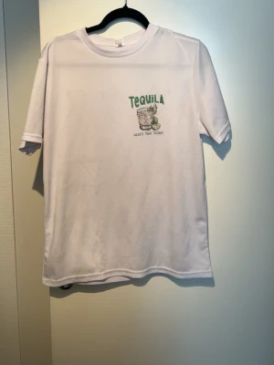 Vit t-shirt med tequila-tryck - Vit t-shirt med grönt tequila-tryck och texten 'Tequila Cheaper Than Therapy' både fram och bak. T-shirten har rund hals och korta ärmar, perfekt för en avslappnad och rolig stil. Sjukt snygg till sommaren och väldigt fräsch 