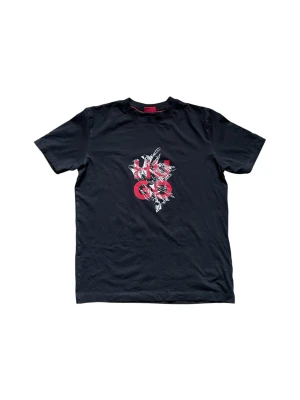 Hugo Boss T-shirt - Svart t-shirt från Hugo Boss med ett grafiskt tryck i rött och vitt på bröstet. Klassisk rund halsringning och korta ärmar. Perfekt för en stilren och modern look. Passa på att fråga om bild eller mått om det behövs! ⭐️