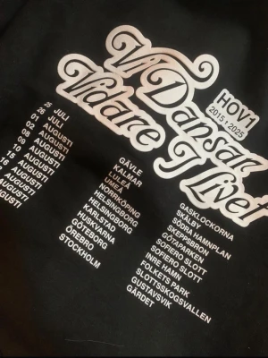 Svart Hov1 hoodie Vi Dansar Vidare - Svart hoodie från Hov1 med stor vit text på ryggen: 'Vi Dansar Vidare I Livet' och turnédatum samt städer. Klassisk huva och magficka. Perfekt för dig som gillar svensk musik och streetwear. Materialet är mjukt och bekvämt, troligen bomullsblandning.
