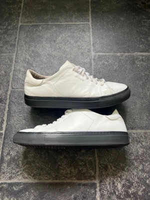 Vita Axel Arigato sneakers med svart sula  - Stilrena vita sneakers från Axel Arigato med svart, chunky sula och vita skosnören. Skorna är låga och har minimalistisk design med diskret logga på sidan. Tillverkade i slätt läder för en clean look som passar till det mesta. Storlek 40 