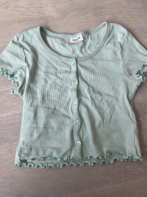 Mintgrön ribbad topp från Kappahl - Söt mintgrön ribbad topp från Kappahl med korta ärmar och knappar framtill. Toppen har volangkant vid ärmslut och nederkant för en extra gullig touch. Perfekt för dig som gillar en trendig och avslappnad stil.