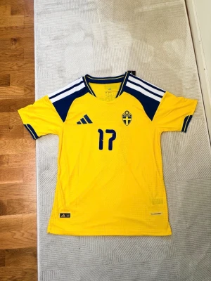 Svensk landslagströja Adidas #17 - Gul svensk landslagströja från Adidas med blå detaljer och vita ränder på axlarna. Nummer 17 och namnet Gyökeres på ryggen. Broderad svensk landslagssköld på bröstet. Tillverkad i lätt och ventilerande material, perfekt för fotboll.