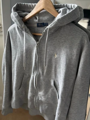 Grå hoodie från Polo Ralph Lauren - Grå zip-hoodie från Polo Ralph Lauren med vit broderad logga på bröstet. Klassisk modell med huva, dragsko och två fickor framtill. Tillverkad i mjuk bomullsblandning för en skön och avslappnad stil.