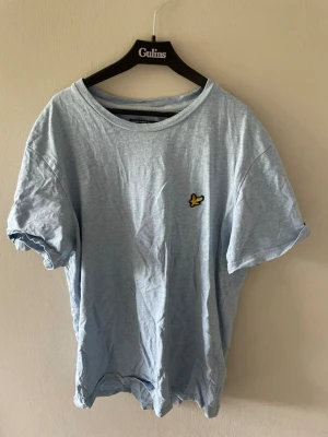 Ljusblå t-shirt från Lyle & Scott - Ljusblå t-shirt från Lyle & Scott med klassisk rund hals och korta ärmar. På bröstet sitter den ikoniska gula örnloggan. Tillverkad i mjuk bomull för en skön och avslappnad känsla.