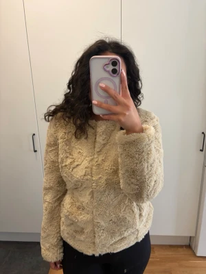 Beige fluffig fuskpälsjacka - Supermjuk och fluffig fuskpälsjacka i beige färg med rund halsringninged knappar framtill. Jackan har en rak passform och är perfekt för att hålla värmen under kyliga dagar. Materialet är fuskpäls som ger en mysig och trendig look.