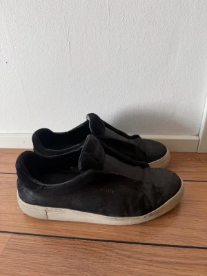 Svarta slip-on sneakers från Axel Arigato - Svarta sneakers från Axel Arigato i slip-on-modell med ovandel i mocka och vit gummisula. Diskret logga i guld på sidan och minimalistisk design utan snörning. Perfekta för en clean och stilren look. Finns box och allt med. En rengöring så är de mycket fina!