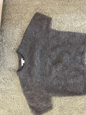 Grå stickad tröja från Gina Tricot - Mysig grå stickad tröja från Gina Tricot med rund halsringning och kortare ärmar. Tröjan har en fluffig och mjuk känsla, perfekt för lager-på-lager och chill dagar. Enkel design som passar till det mesta.